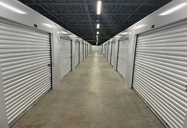 Interior hallway with white roll-up storage unit doors lining both sides, fluorescent ceiling lights, concrete floor.